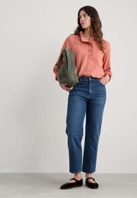 Koraalrode blouse met ruffles bij de hals, blauwe recht gesneden jeans en donkerbruine platte schoenen; model houdt een grijsgroene tas vast.