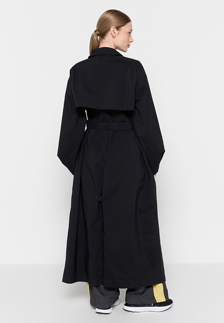 Czarny płaszcz typu trench coat o luźnym kroju, z długimi rękawami i paskiem. Posiada przyciętą nakładkę z tyłu i gładki, lekki materiał.