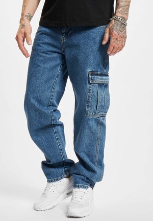 Mężczyzna ubrany w niebieskie dżinsowe spodnie cargo z boczną kieszenią, białe trampki, czarną koszulę i tatuaże na rękach i ramionach na białym tle.