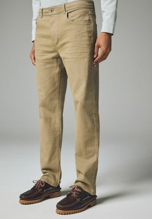 Beige cotton chinos with a slim fit, featuring a classic five-pocket design and a subtle texture, paired with dark brown boat shoes.