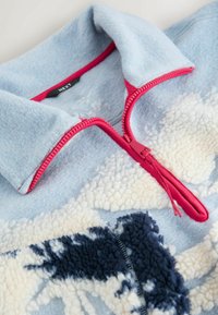 Fliisist pullover, millel on helesinine keha, roosa tõmblukk ning tekstureeritud valged ja tumesinised mustrid. Omab kõrget kraed ja pehmeid kangaid.