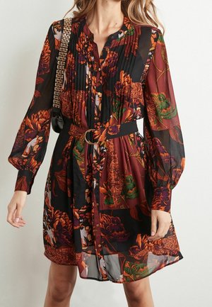 Frau trägt ein langärmliges, transparentes Blumenmuster-Kleid in Schwarz, Dunkelrot, Orange und Grün mit einem Taillengürtel, trägt eine schwarze Tasche mit goldener Kettenriemen.