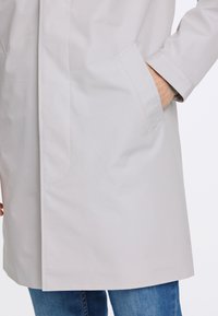 Impermeabile grigio chiaro con una texture liscia, dotato di tasche laterali, taglio dritto e accenti di cucitura ben visibili. Indossato su jeans blu.