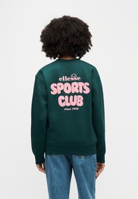 Άτομο με σγουρά μαλλιά φοράει σκούρο πράσινο φούτερ με ροζ κείμενο "ellesse SPORTS CLUB since 1959" στην πλάτη και μπλε τζιν.