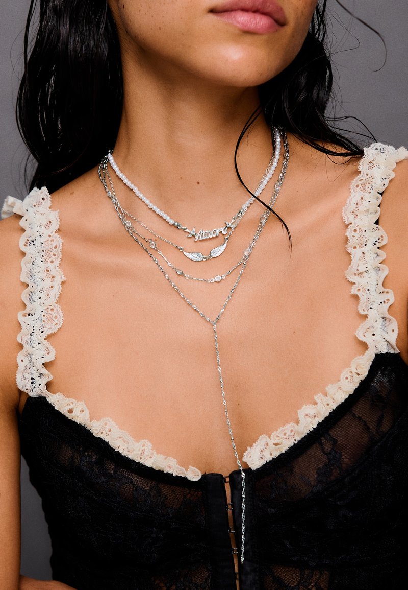 Femme portant un haut en dentelle noire avec des bretelles volantées crème et des colliers en argent superposés, dont un avec le mot "Amore".