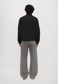 Black lightweight jacket with a stand collar, shown from the back, paired with loose grey denim pants with a faded texture and wide legs.