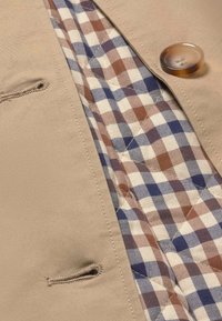 Chaqueta de tela exterior beige con cierre de botón; forro a cuadros en marrón, azul y crema con textura acolchada; cuenta con un pequeño bolsillo lateral.