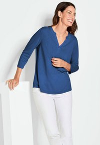 Blaues, langärmeliges Oberteil mit V-Ausschnitt-Design. Sanfte Textur, lockerer Schnitt und dezentes Muster. Kombiniert mit weißen Hosen, minimalistischem Hintergrund.