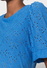 Zblízka pohled na osobu, která nosí jasně modrou blůzu z očkované krajky s květinovými vzory a nařasenými krátkými rukávy.