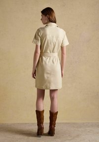 Sieviete, tērpusies bēša krāsas īsām piedurknēm kleitā ar jostu un valkājot brūnas rietumu stila papēžu zābakus, stāvot pretī teksturētai bēša krāsas sienai.