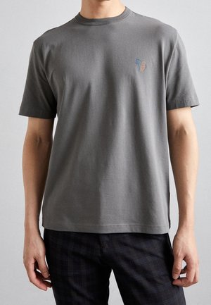 Uomo che indossa una maglietta a maniche corte grigia semplice con un piccolo logo colorato di un cane sul petto a sinistra e pantaloni scuri a quadri.