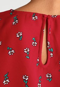 Tissu rouge avec des imprimés de fleurs blanches et des tiges vertes. Dispose d'une ouverture en forme de trou à l'arrière, fermée par un bouton. Texture douce.