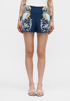 Γυναίκα που φορά σορτς σε μπλε ναυτικό χρώμα με χρυσά και λευκά μπαρόκ floral σχέδια, λευκό αμάνικο τοπ, λευκά σανδάλια και εμφανή τατουάζ στα χέρια.