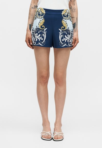 Femme portant un short bleu marine avec des motifs floraux baroques dorés et blancs, un top blanc sans manches, des sandales blanches et des tatouages visibles sur les bras.