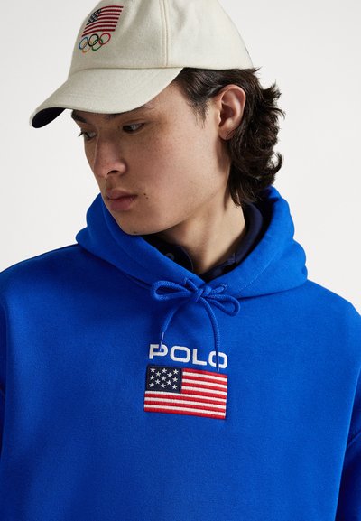Blå hettegenser med snøring og "POLO"-bokstaver, samt et amerikansk flaggmerke. Beige caps med OL-ringene og flaggemblem.