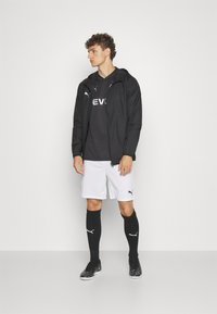 Zwarte windjack, zwart T-shirt met logo, witte shorts, zwarte kniekousen en zwarte voetbalschoenen. Model staat zijwaarts.