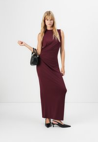 Sieviete ar gariem gaišiem matiem, tērpusies bez piedurknēm burgundijas krāsas maxi kleitā, melnās slingback kurpēs un turot melnu rokassomu uz baltas fona.