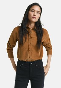 Brown corduroy button-up shirt with long sleeves, featuring a collar and subtle texture, paired with dark jeans.