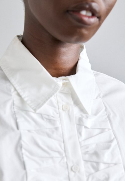 Chemise blanche avec un col pointu, présentant des détails en volants sur le devant et une fermeture à bouton standard. Tissu à texture lisse.
