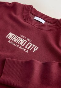 Bordowy sweter z prążkowanym kołnierzem, z białym napisem "NAKANO CITY" i "TOKYO" w wyrazistym kroju, gładka tekstura i luźny krój.