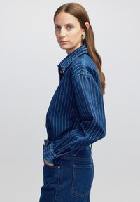 Jeansjacke mit vertikalen weißen Streifen, Knopfleiste und spitzem Kragen. Der Stoff ist weich und hat einen leicht verblassten Blauton.