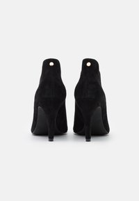 Botas de tacón alto de ante negro con un diseño elegante, presentan una puntera redondeada y un detalle de botón dorado metálico en la parte trasera.
