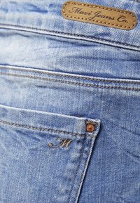 Vaaleansiniset farkut, joissa on takataskussa ruskea nahkainen brändilogo "Marie Jeans Co." ja brodeerattu kirjain "M" taskun kulmassa.