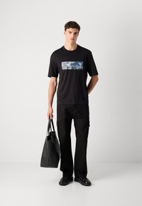 Ung mand iført sort t-shirt med grafik og bukser, holder en sort totebag, står foran en ensfarvet hvid baggrund.