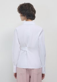 Camicia bianca a maniche lunghe con colletto a punta e dettaglio in crêpe sul retro, indossata con pantaloni rosa pallido. Texture liscia, vestibilità sartoriale.