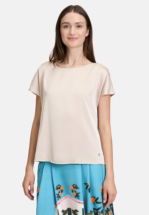 Femme portant un chemisier crème à manches courtes et une jupe bleue avec des motifs de fruits orange sur un fond gris clair.