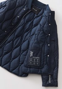 Giacca trapuntata blu navy con colletto a coste, chiusura con zip e bottoni a pressione. Presenta un'etichetta interna con icone e dettagli sui materiali.