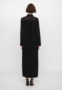 Vestido maxi negro con mangas largas de tul, cuello con botones y un corte holgado. La parte posterior presenta un sutil detalle con pliegues. Combinado con tacones negros.