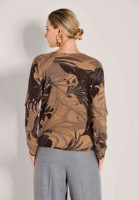 Vrouw met blond haar in een knot, gekleed in gouden oorbellen, een bruine trui met een zwart bladerenpatroon, en grijze broek, staande tegen een beige achtergrond.