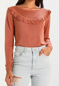 Haut à manches longues couleur rouille avec un col rond orné d'un volant. Assorti à un jean taille haute bleu clair déchiré.