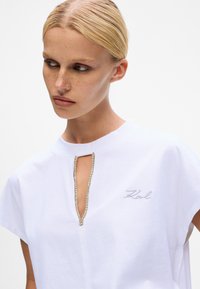 Weißes Baumwoll-T-Shirt mit kurzen Ärmeln, mit einem Schlüssel-Ausschnitt, der mit einem silberfarbenen Rand und gesticktem "Karl"-Detail verziert ist.