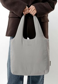 Szara torba wielokrotnego użytku o gładkiej, błyszczącej fakturze i dwóch mocnych uchwytach, trzymana przez osobę w brązowym płaszczu i dżinsach.