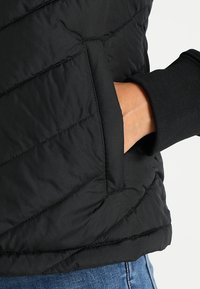 Schwarze isolierte Weste mit einem rautenförmigen Steppmuster, Seitentasche und gerippten Bündchen, aus glattem, leichtem synthetischem Material hergestellt.