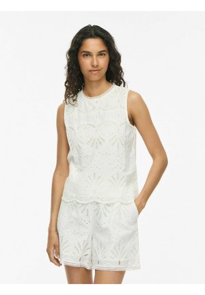 Femme aux cheveux bouclés portant un haut blanc sans manches en dentelle ajourée et un short assorti, debout avec une main dans la poche.