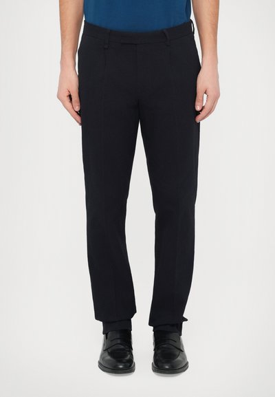 Pantalon noir sur mesure en tissu texturé, taille plate et passants pour ceinture, associé à des chaussures noires brillantes, posé sur un fond neutre.