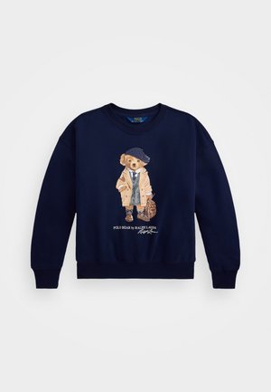 Sudadera azul marino con un oso bordado vestido con un abrigo beige y un sombrero azul, con una bolsa decorativa y texto debajo del gráfico.