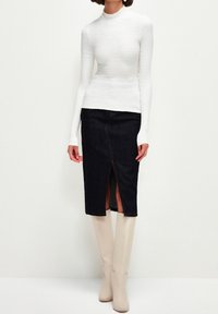 Top bianco a maniche lunghe con texture, abbinato a una gonna in denim scuro a vita alta con spacco frontale, completato da stivali beige alti fino al ginocchio.