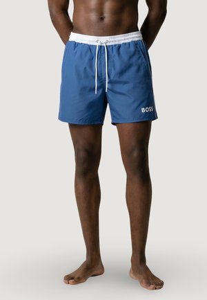 Torso et jambes d'un homme portant un short de bain bleu BOSS avec une ceinture blanche et un cordon de serrage, debout pieds nus devant un fond uni.