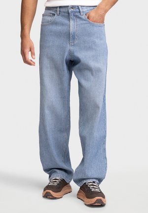 Homme portant un jean bleu clair ample, une main dans la poche, et des baskets marron avec des lacets blancs sur fond clair.