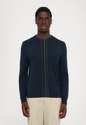 Mann trägt einen marineblauen Pullover mit langen Ärmeln und bunten vertikalen Streifen in der Mitte, kombiniert mit beigen Hosen, und steht vor einem weißen Hintergrund.