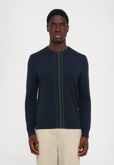 Homme portant un pull à manches longues bleu marine avec des rayures verticales colorées au centre, associé à un pantalon beige, se tenant debout devant un fond blanc.