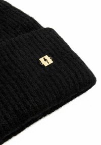 Czarna dziana czapka z prążkowanym wzorem, z złotą kwadratową metką z wyciętym wzorem. Miękki materiał wełniany, zbliżenie.