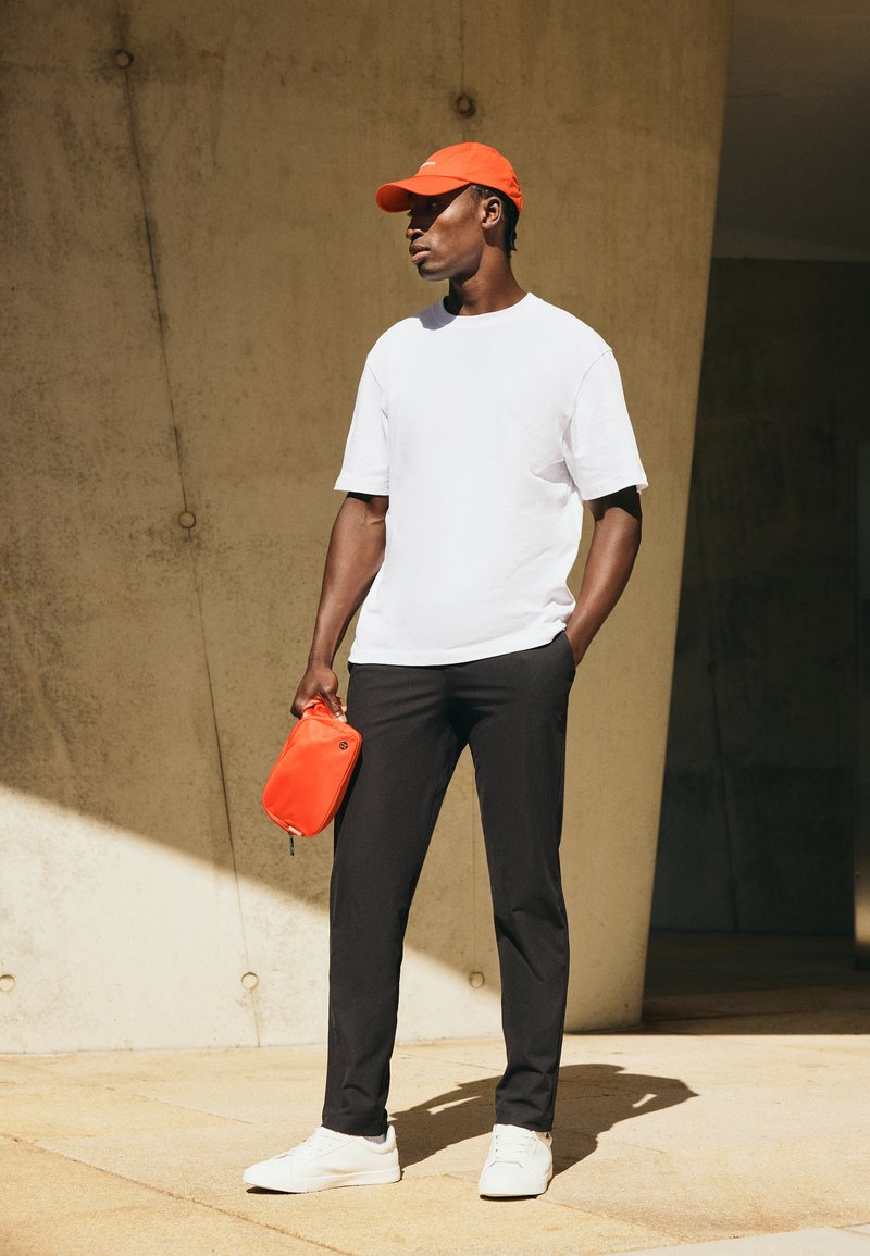 Man draagt een wit T-shirt, zwarte broek, witte sneakers en een oranje pet, houdt een oranje etui vast en staat tegen een beige betonnen muur.