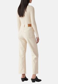 Jeans beige chiari a vita alta con gambe dritte, tessuto di cotone testurizzato e patch sulla vita posteriore. Mules con tacco neri.