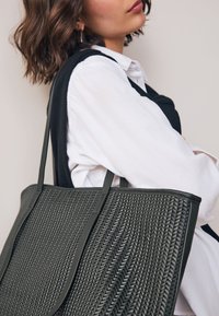 Sac fourre-tout vert tissé avec motif texturé et poignées solides, présentant une forme rectangulaire plate et un design minimaliste.