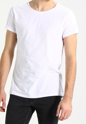 Mann trägt schlichtes weißes T-Shirt mit kurzen Ärmeln und schwarzen Jeans, steht vor weißem Hintergrund.